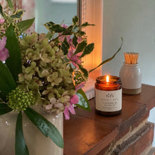 Load image into Gallery viewer, The Embers and Roots gingerbread christmas scented soy candle on a brick mantelpiece in a cottage next to a vintage jar of flowers and a match striker