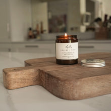 Load image into Gallery viewer, An amber glass jar candle with a white label reading 'Embers + Roots' and 'Gingerbread,' lit and placed on a wooden cutting board in a softly blurred kitchen setting. The silver metal lid rests beside the jar.