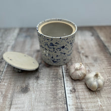 Load image into Gallery viewer, A handmade garlic pot in a splatter ware glaze on a wooden table with its lid to one side and a couple of garlic bulbs alongside it 