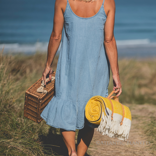 A woman strolling through sand dunes with her picnic basket and her yellow herringbone picnic blanket from Atlantic Blankets